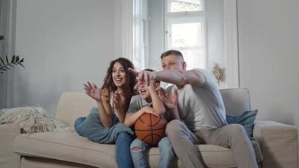 Jonge familie op de bank in de kamer. Ze kijken TV en juichen voor sport spelletjes. — Stockvideo