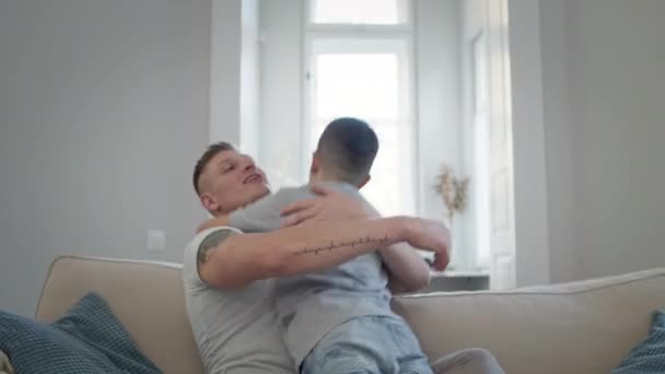 Hombre joven sentado en el sofá en el apartamento. Su hijo corre hacia él. — Vídeos de Stock