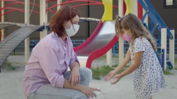 Giovane madre e bambina per strada in maschere mediche per proteggere contro le infezioni virali. Mamma spruzza antisettico sulle mani delle ragazze. — Video Stock