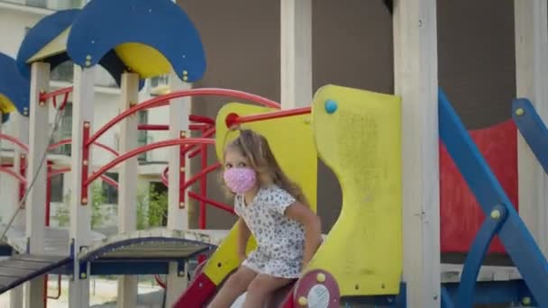 Mała dziewczynka w masce medycznej na placu zabaw. Schodzi ze zjeżdżalni.. — Wideo stockowe