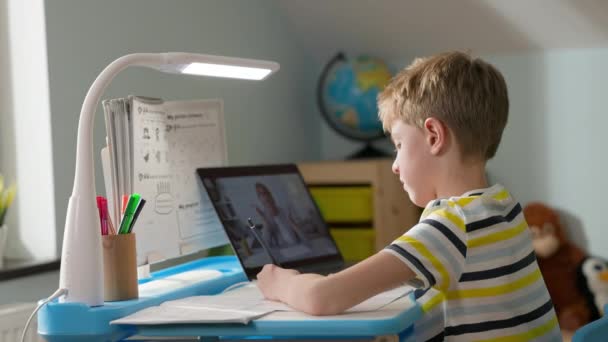 Niño sentado en el portátil en casa. Lección en línea. Niño escribe en el cuaderno mientras mira la pantalla del ordenador portátil. — Vídeos de Stock