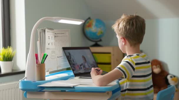 Chłopiec siedzi na laptopie w domu. Lekcja online. Chłopiec pisze w notatniku. — Wideo stockowe