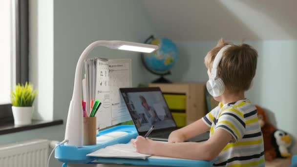 Chłopiec siedzi na laptopie w domu. Lekcja online. Dziecko w słuchawkach słucha lekcji online i pisze w notatniku. Uczeń podnosi rękę, aby uzyskać pozwolenie na wypowiedź. — Wideo stockowe