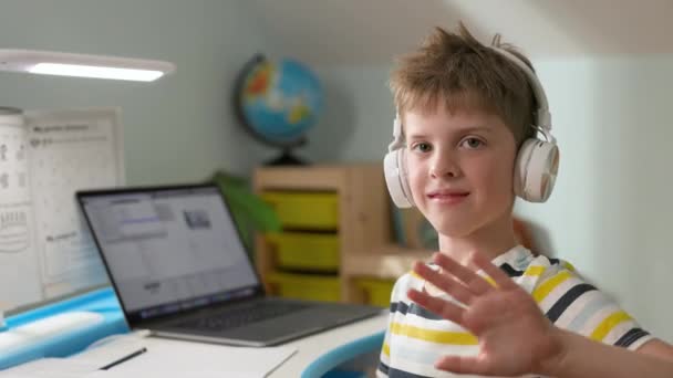 Blond pojke av sju år i hörlurar ler. Pojken sitter hemma på laptop. Han viftar med handen i hälsning. — Stockvideo