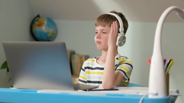 Chlapec sedí doma u laptopu. Vzdálené učení. Sedmiletý chlapec ve sluchátkách zvedá ruku, aby odpověděl. — Stock video