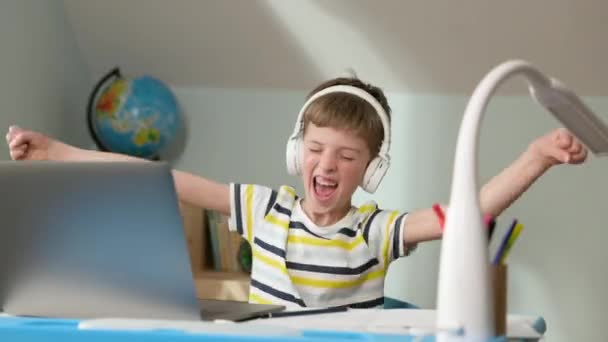 Chłopiec siedzi na laptopie w domu. Lekcja online. Ma słuchawki. Coś sprawia, że chłopiec jest bardzo szczęśliwy i podnosi ręce. — Wideo stockowe