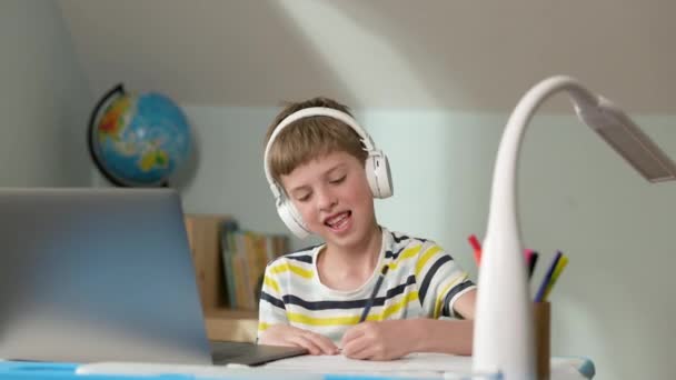 Chłopiec siedzi na laptopie w domu. Ma słuchawki. Pisze w notatniku. Coś sprawia, że chłopiec jest bardzo szczęśliwy i podnosi rękę. — Wideo stockowe