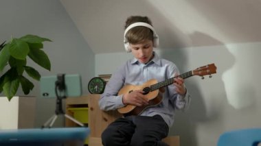 10 yaşındaki çocuk elinde gitarla bebek odasında oturuyordu. Telefonun önünde, kulaklıkla gitar çalıyor. Çevrimiçi Ders.