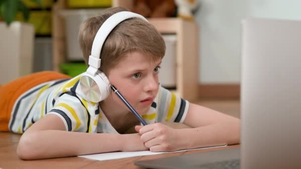Pojke liggandes på golvet framför laptop. Söt pojke i sju år i hörlurar. — Stockvideo