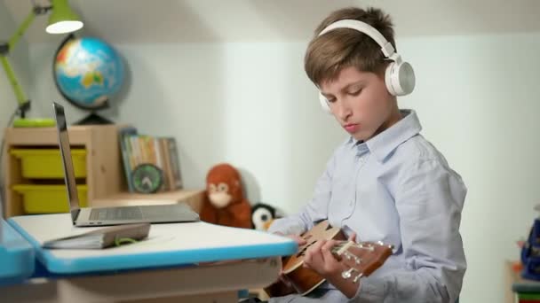 Junge sitzt zu Hause am Laptop. Junge im Kopfhörer spielt Gitarre. — Stockvideo