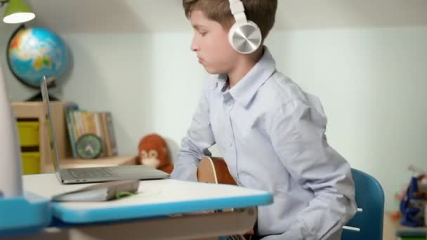 Niño sentado en el portátil en casa. Niño en auriculares toca la guitarra. — Vídeos de Stock