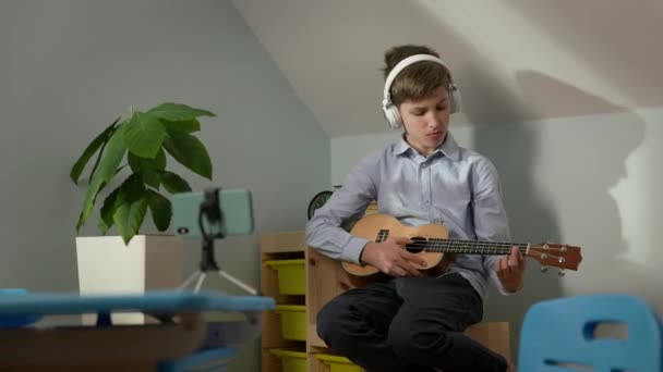 Ragazzo di dieci anni seduto nella nursery con la chitarra in mano. Suona la chitarra con le cuffie davanti al telefono. Lezioni online. — Video Stock