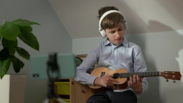 Dziesięciolatek siedzący w pokoju dziecinnym z gitarą w rękach. Gra na gitarze w słuchawkach przed telefonem. Lekcja internetowa. — Wideo stockowe