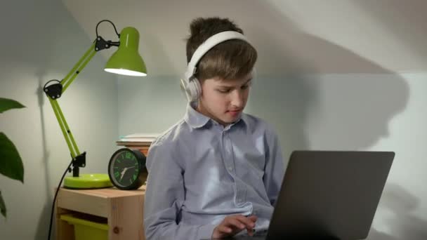 Um rapaz de 10 anos sentado no berçário com um portátil na mão. Rapaz move a cabeça para a batida da música. — Vídeo de Stock