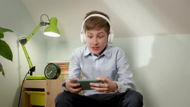 Dziesięciolatek siedzi w pokoju z telefonem w rękach. Słuchawki na głowie. — Wideo stockowe