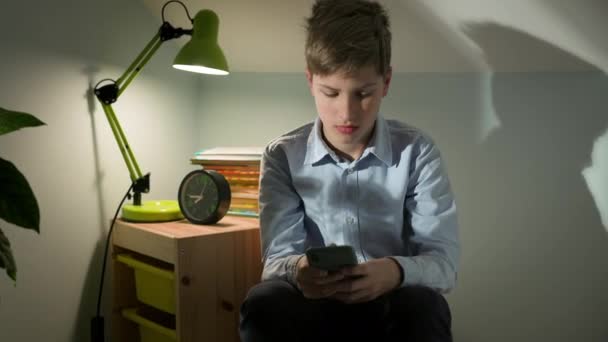 Un niño de diez años está sentado en su habitación con el teléfono en las manos. Auriculares en su cabeza. Habitación está iluminada por la lámpara. — Vídeos de Stock