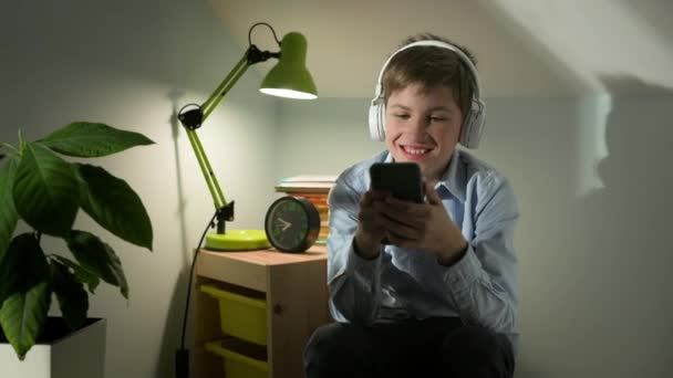 Deset let starý chlapec sedí ve svém pokoji s telefonem ve svých rukou. Sluchátka na hlavě. — Stock video