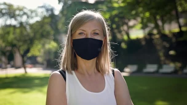 Mujer envejecida de pie en el parque en el soleado día de verano. En su máscara facial para protegerse contra infecciones virales. — Vídeo de stock
