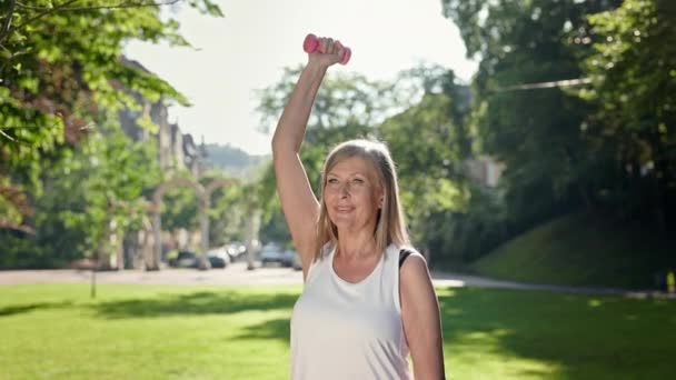 Verouderde vrouw gaat sporten in het park. Ze heft haar arm op met duimbellen en haalt diep adem.. — Stockvideo