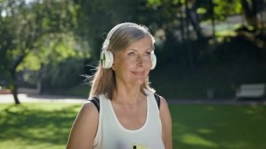 Yaşlı kadın kulaklıklarla parkta koşuyor. Ellerinde telefonu tutuyor..