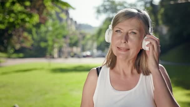 Grijze vrouw in het park luistert naar muziek op koptelefoon. In haar handen De telefoon. — Stockvideo
