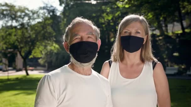 Mężczyzna i kobieta w podeszłym wieku stoją w parku. Są w maskach medycznych. Wspaniały słoneczny dzień dla fitness. — Wideo stockowe