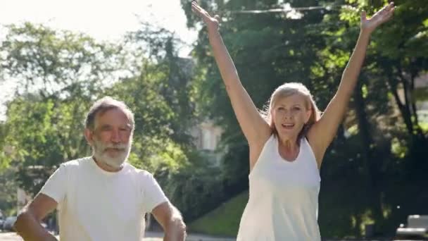 Oude man en vrouw die sporten in het park. Ze rennen vrolijk glimlachend en steken hun handen op. — Stockvideo