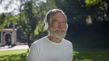 Yaşlı adam kulaklıklarla parkta koşuyor. Güneşli yaz gününde Sabah Koşusu.