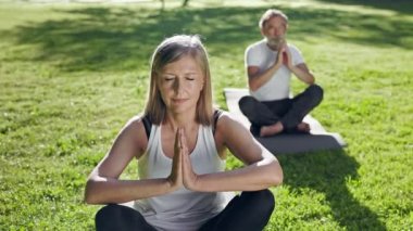 Yaşlı Adam ve Kadın Park 'ta Yoga Yapıyor. Yerdeki halılara otururlar. Onların gözleri kapalıdır. Kadının Arkasındaki Adam.