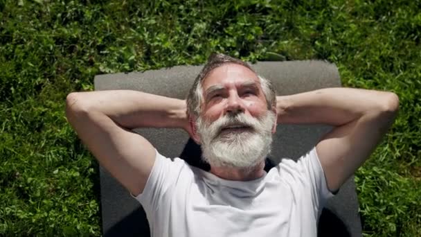 Starší muž leží na trávě na sportovní podložce. Dívá se na oblohu. — Stock video