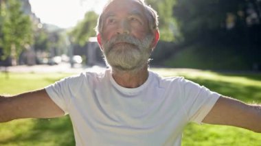 Yaşlı gri saçlı adam parkta spor yapıyor. Ellerini kaldırır ve indirir..
