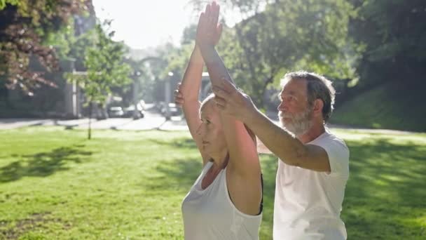 Пожилой мужчина и женщина с седыми волосами занимаются фитнесом в парке. Мужчина исправляет женские руки. — стоковое видео