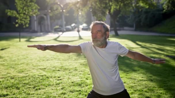Oudere grijze harige man doet fitness in het park. Hij houdt zijn armen uitgestrekt in verschillende richtingen.. — Stockvideo