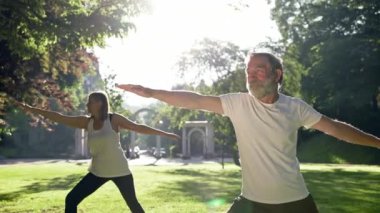 Yaşlı adam ve gri saçlı kadın parkta spor yapıyor. Kollarını açık tutarlar..