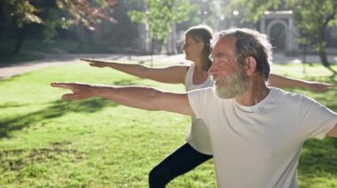 Yaşlı adam ve gri saçlı kadın parkta spor yapıyor. Kollarını açık tutarlar..