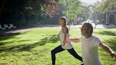 Yaşlı adam ve gri saçlı kadın parkta spor yapıyor. Bir ayağı önde durur ve avuç içlerine dokunarak ellerini kaldırırlar..