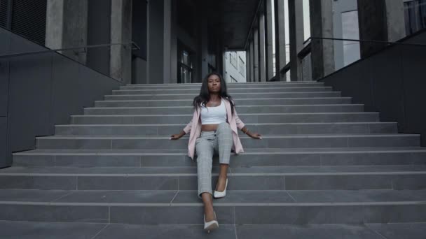 Joven chica negra sentada en las escaleras del edificio. Ella está elegantemente vestida y con tacones altos. — Vídeo de stock