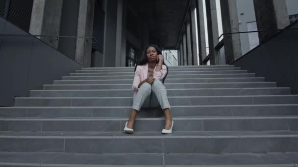 Молодая чёрная девушка, сидящая на лестнице здания. Она стильно одета. — стоковое видео