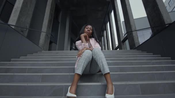 Jeune fille noire assise sur les escaliers du bâtiment. Elle est élégamment habillée. Elle touche son menton avec sa main. — Video