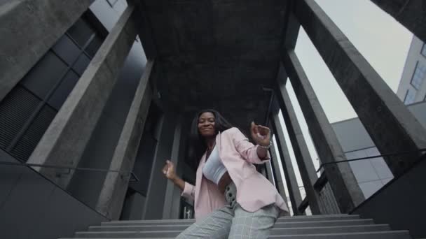 Jeune fille noire dansant danse moderne sur les escaliers. Elle porte des vêtements d'affaires avec des talons hauts. Vue du bas. — Video
