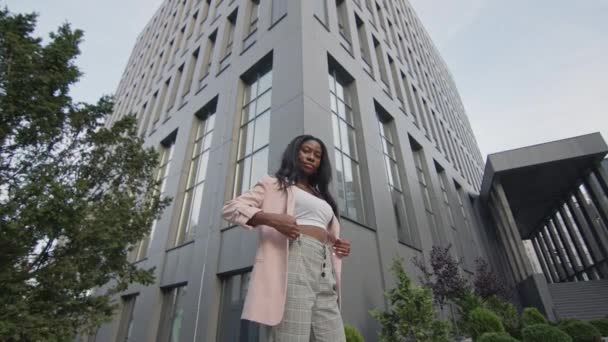 Rapariga negra muito jovem fica na rua. Menina bonita com cabelo comprido. Atrás de seu alto prédio de escritórios. — Vídeo de Stock