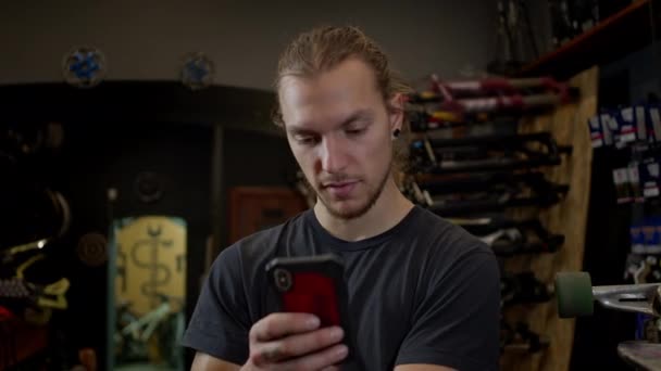 Junger Mann mit Handy in der Hand steht in Fahrradwerkstatt Er benutzt aktiv das Telefon. Überrascht zieht er die Augenbrauen hoch. — Stockvideo