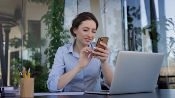 Mladá dívka pracující v kanceláři. Sedí u stolu a drží mobilní telefon ve svých rukou. Girl Smiles. — Stock video