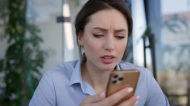 Ofisteki genç bayan işçi. Masada oturuyor ve cep telefonunu avucunun içinde tutuyor. Sinirli bir şekilde parmağını telefon ekranına bastırıyor. Çalışmıyor gibi görünüyor..