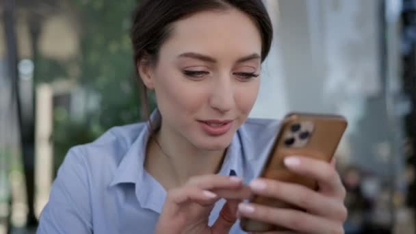 Porträtt av en ung flicka på kontoret. Hon sitter vid bordet och håller mobiltelefonen i händerna. Flickan skriver meddelande och ler. Närbild. — Stockvideo