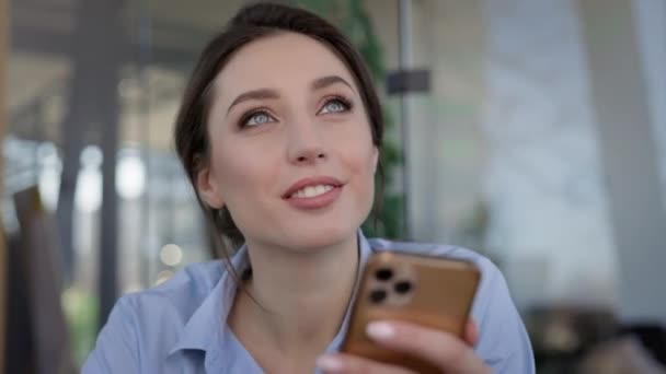 Junge Arbeiterinnen im Büro. Sie sitzt am Tisch und hält das Handy in den Händen. Mädchen lächelt. Sie denkt einen Moment lang nach. — Stockvideo