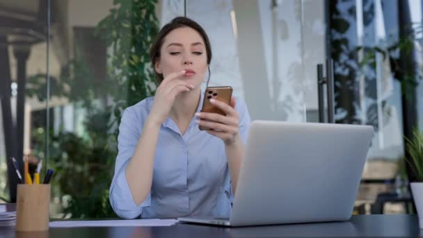 Mladá dívka pracující v kanceláři. Sedí u stolu a drží mobilní telefon ve svých rukou. Myslí si to. Dívka znepokojená korespondencí. — Stock video