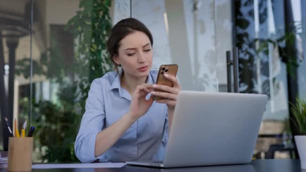 Fiatal lány munkás az irodában. Az asztalnál ül és mobiltelefont tart a kezében.. — Stock videók