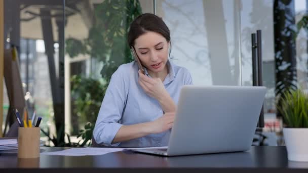 Mladá dívka pracující v kanceláři. Pěkná bruneta sedí u laptopu a pracuje. Pracuje s dokumenty. Drží v ruce telefon. — Stock video