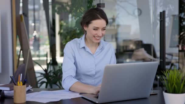 Mladá dívka pracující v kanceláři. Pěkná bruneta sedí u laptopu. Zamává někomu, kdo se dívá na monitor notebooku. — Stock video